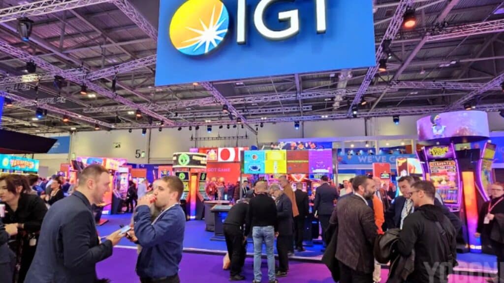 A wide view of a busy trade show floor under a large blue "IGT" sign. Numerous attendees in business attire are networking and walking between various gaming booths and displays in a large convention hall.