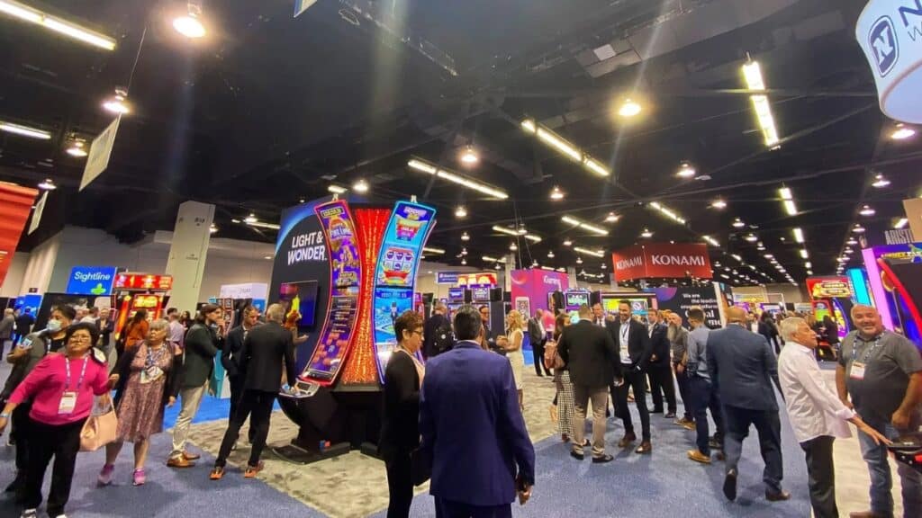 A perspective shot of a crowded convention hall featuring various gaming exhibits. A prominent "Light & Wonder" display stands in the foreground, surrounded by attendees, with "Konami" and "iGaming" booths visible in the distance under bright ceiling lights.