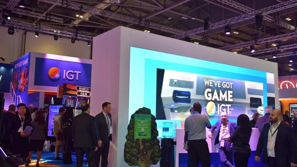 A view of the IGT booth at a trade show, featuring a large digital screen with the slogan "WE'VE GOT GAME." Attendees are seen walking past the booth and conversing, with purple and blue event lighting illuminating the structure.