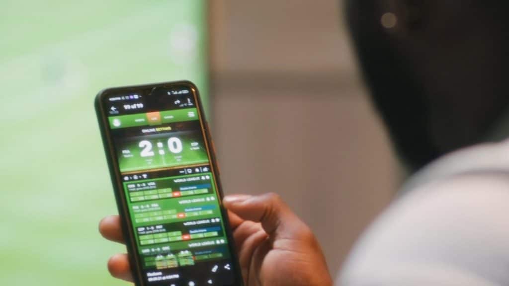 A close-up, first-person view of a person holding a smartphone displaying a live sports betting application. The screen shows real-time scores and odds for various matches, illustrating the digital landscape of texas sports betting through mobile platforms.