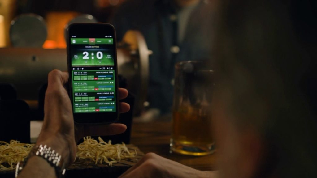 A first-person view of a person using a mobile sports betting app in a dimly lit bar setting. The screen shows live odds and scores, illustrating the consumer experience that would be impacted by the proposed Colorado Sports Betting Reform Bill SB 26 131.