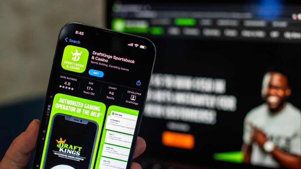 A smartphone displaying the DraftKings logo in the foreground, set against a blurry background of financial stock market candles, representing the expansion of DraftKings Prediction Markets.