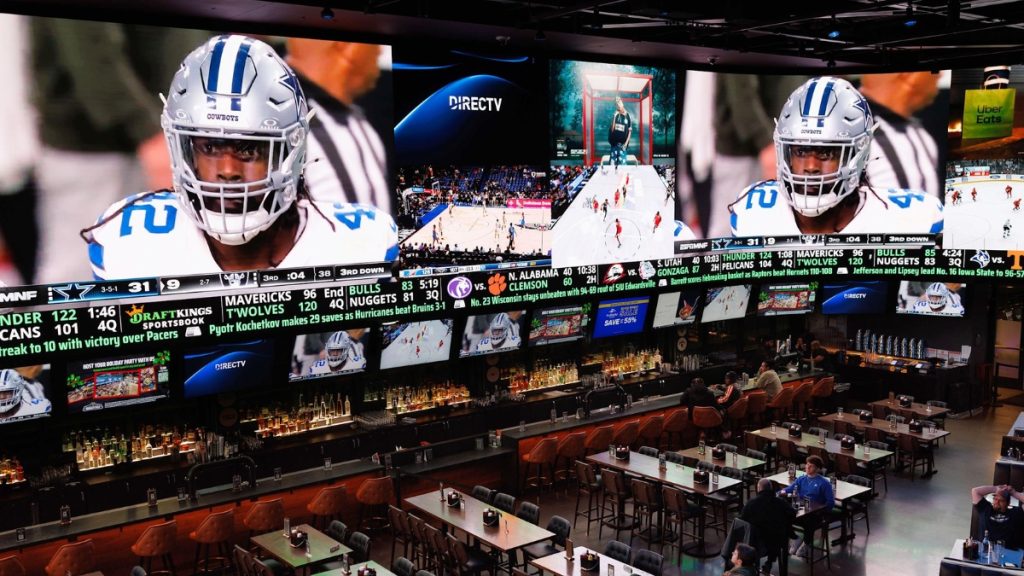 The interior of the DraftKings Sportsbook Wrigley location, showing a massive wall of video screens broadcasting live sports, a long bar, and restaurant seating areas.