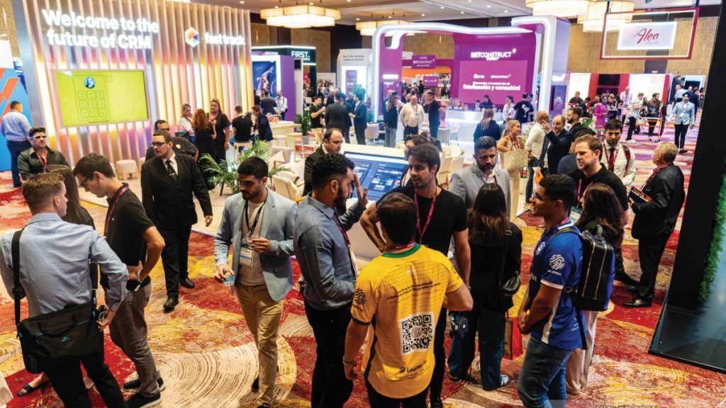 A wide-angle view of a bustling trade show floor, similar to the setting of SBC Summit Americas, featuring the large, pink, circular BetConstruct booth, Golden Race branding, and diverse attendees networking.