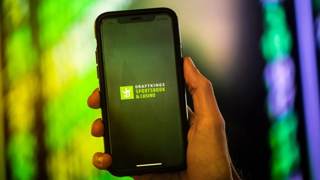 A close-up of a hand holding a smartphone displaying the DraftKings Sportsbook & Casino app logo against a blurred neon background, highlighting a major operator authorized by the Massachusetts Sports Betting Bill.