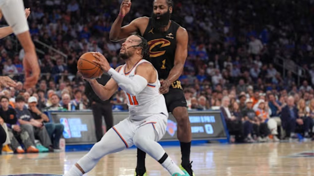Action shot from a professional basketball game, used to promote Knicks vs cavs game 4. New York Knicks point guard Jalen Brunson is driving aggressively through the paint toward the basket, tightly guarded by multiple Cleveland Cavaliers defenders in their white home uniforms under the bright arena lights.