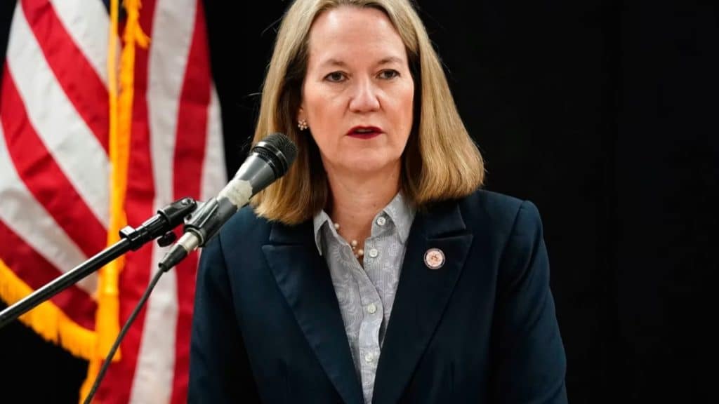 A portrait of a woman with shoulder-length blonde hair, identified as co-founder Luana Lopes Lara, wearing a navy blazer and patterned blouse while speaking at a podium with a microphone. An American flag is visible in the background, symbolizing the Kalshi prediction market's focus on federal regulation and U.S. event contracts.