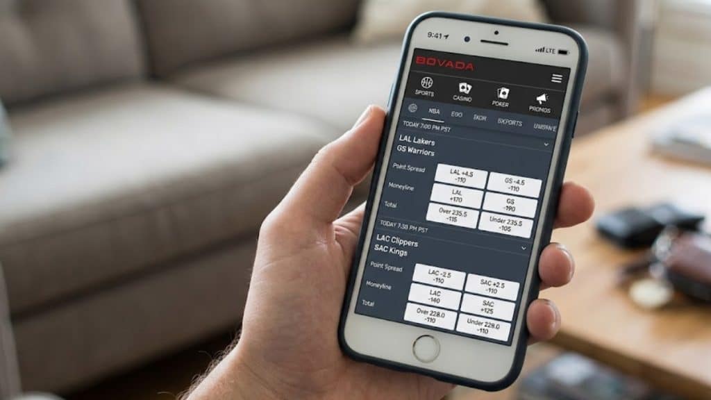 A close-up view of a hand holding a smartphone displaying the Bovada mobile sportsbook interface, showing live point spreads and moneyline odds for a basketball game between the Lakers and the Warriors.