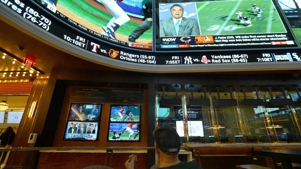 The interior of a modern sportsbook lounge featuring a massive electronic ticker showing live sports scores, betting odds, and upcoming game times for teams like the Red Sox and Orioles. In the foreground, a patron watches multiple TV screens broadcasting live games, a scene central to the debate over whether sports betting is still legal under the Massachusetts Sports Betting Bill June 2026.