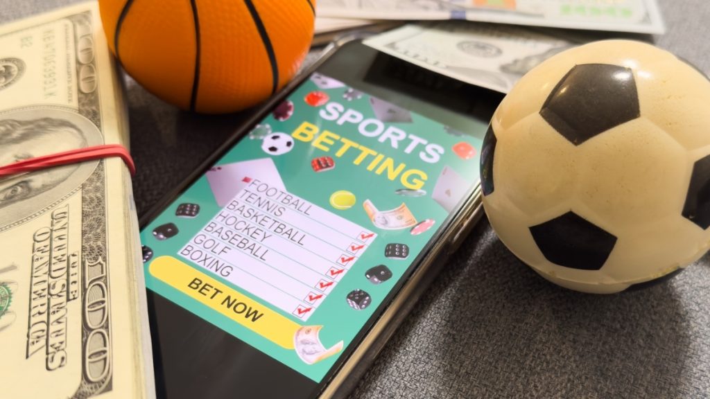 A smartphone showing an active digital sports wager checklist alongside stacks of cash, a mini basketball, and a soccer ball, representing the potential impact of a mississippi online sports betting bill.