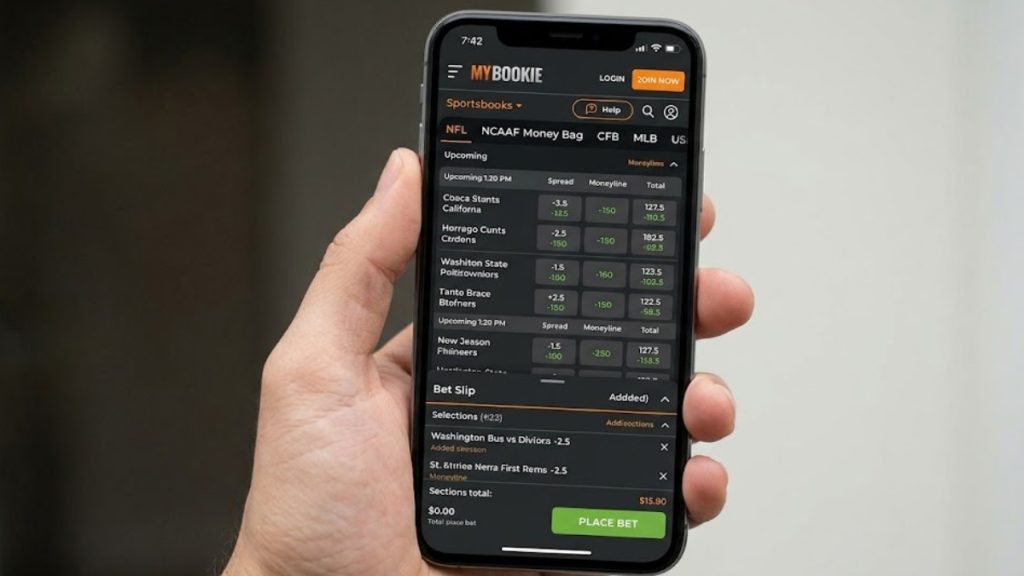 A close-up of a hand holding an iPhone displaying the MyBookie mobile sportsbook interface, relevant for a MyBookie vs BetUS mobile review. The screen shows upcoming NFL betting lines, point spreads, and money lines, with an active bet slip open at the bottom.