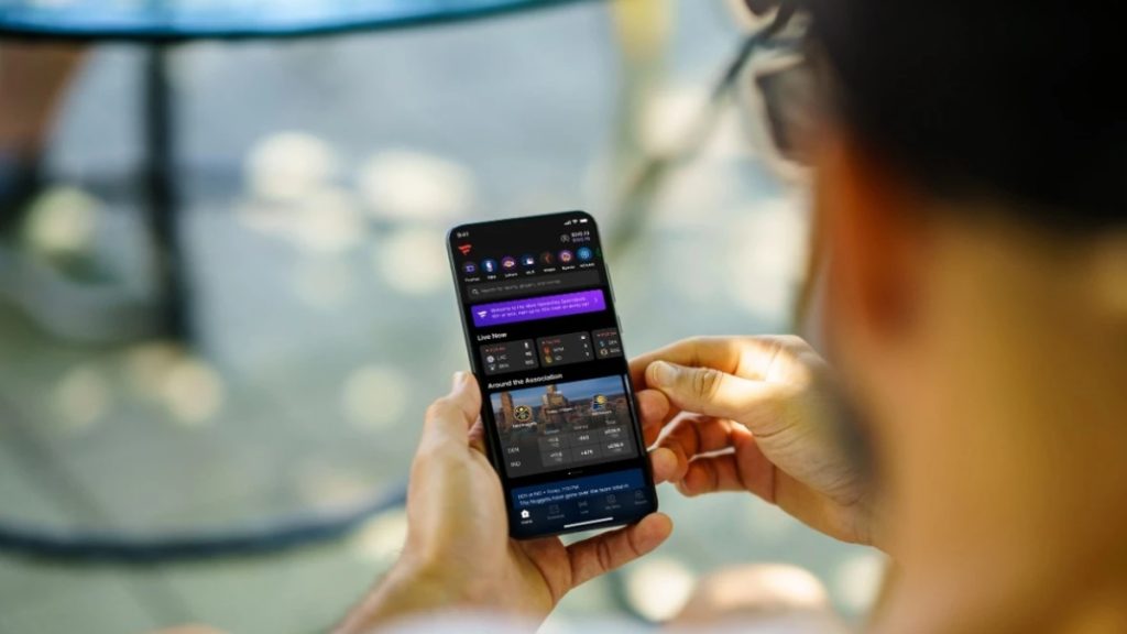 A person holding a smartphone outdoors, viewing a mobile sports betting app interface displaying upcoming live basketball game odds. This mobile wagering visualization represents the digital expansion being discussed within a potential Wisconsin Sports Betting Bill.