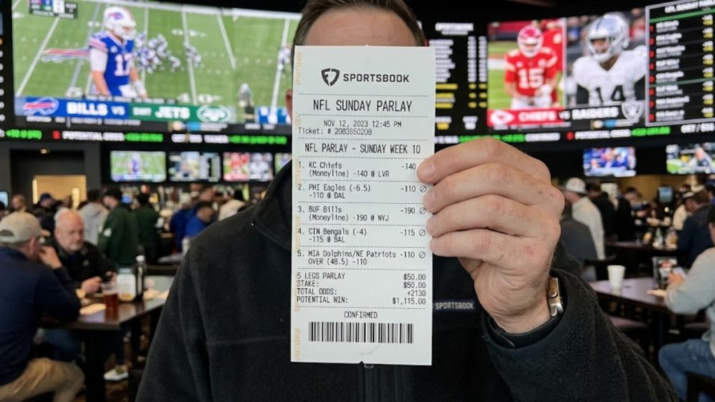 A person holding up a physical sports betting ticket showing NFL Parlays inside a crowded sportsbook with TVs playing football games in the background